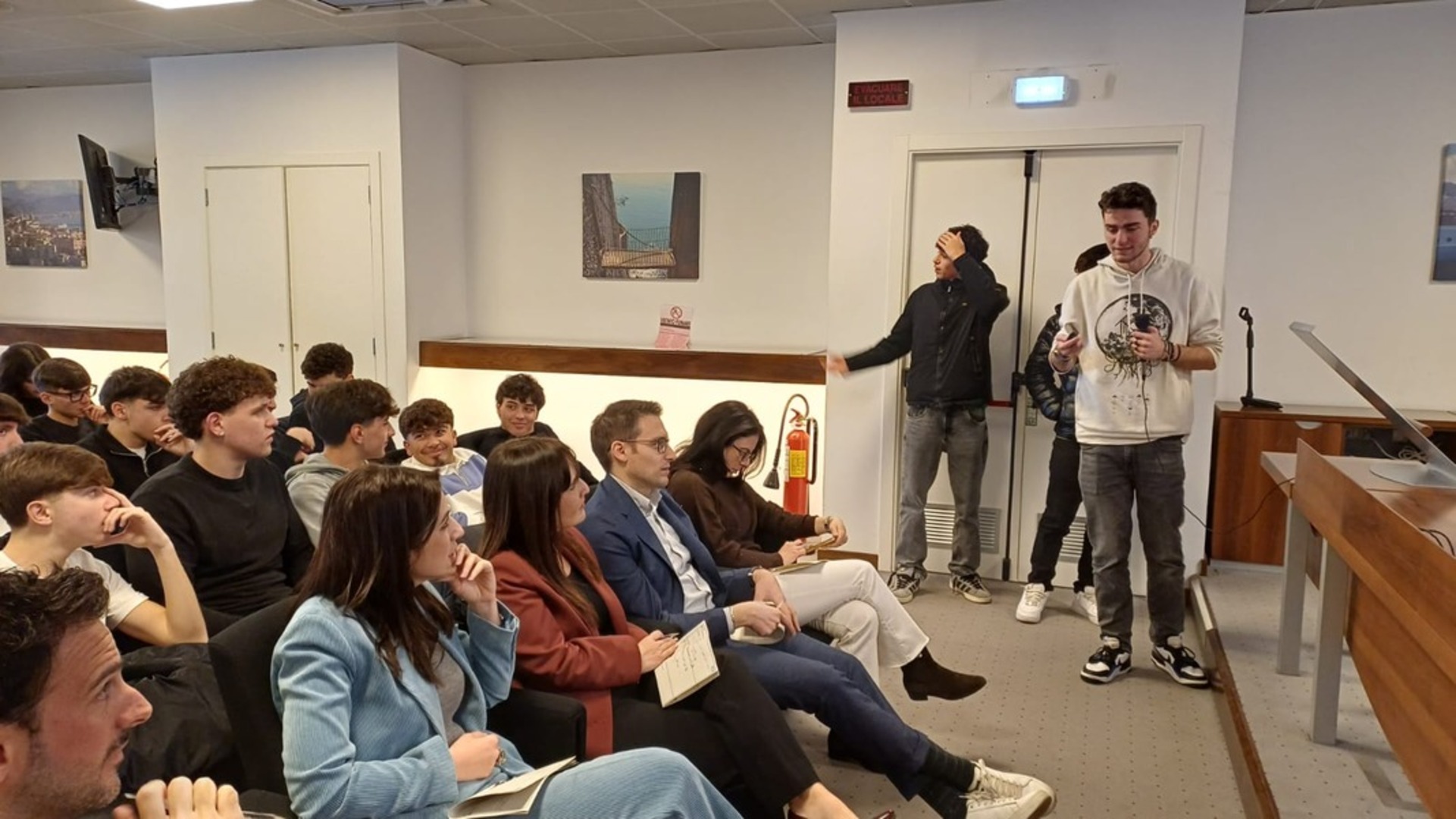 Una platea gremita di giovani studenti e professionisti seduti ascolta con attenzione un ragazzo sul palco dell'Hub Rete di Salerno che presenta il suo progetto d'impresa.